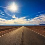 A long, straight road stretches across a dry desert landscape under a bright blue sky and shining sun.
