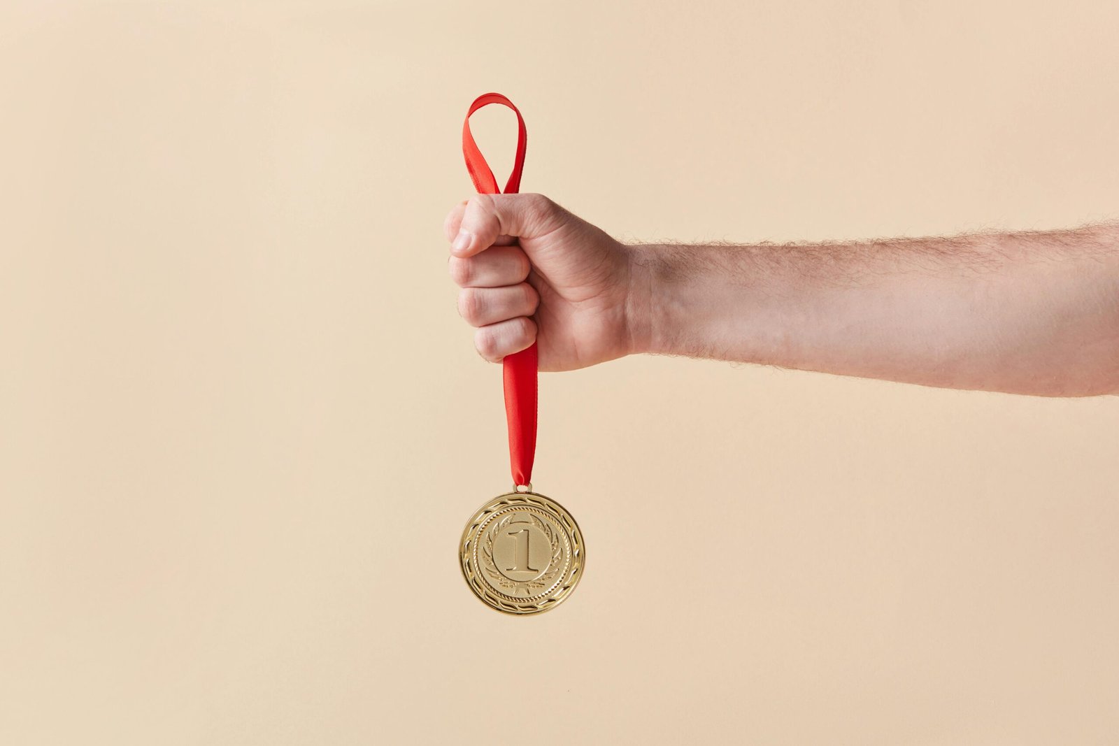 A hand proudly holds a gold medal with a red ribbon on a beige background.